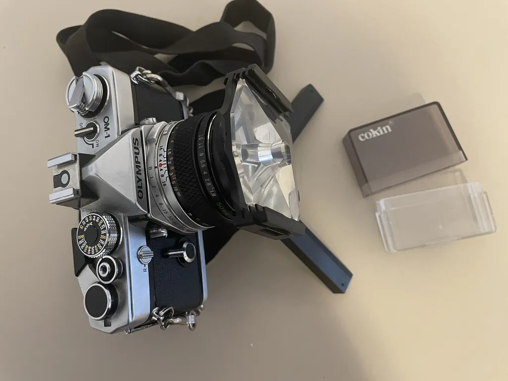 a black and silver metal camera on a tiny tripod. On the front of the lens is a black piece of plastic with ridged slots on either side. a thick, angular piece of plastic with a hole in the center is slotted in