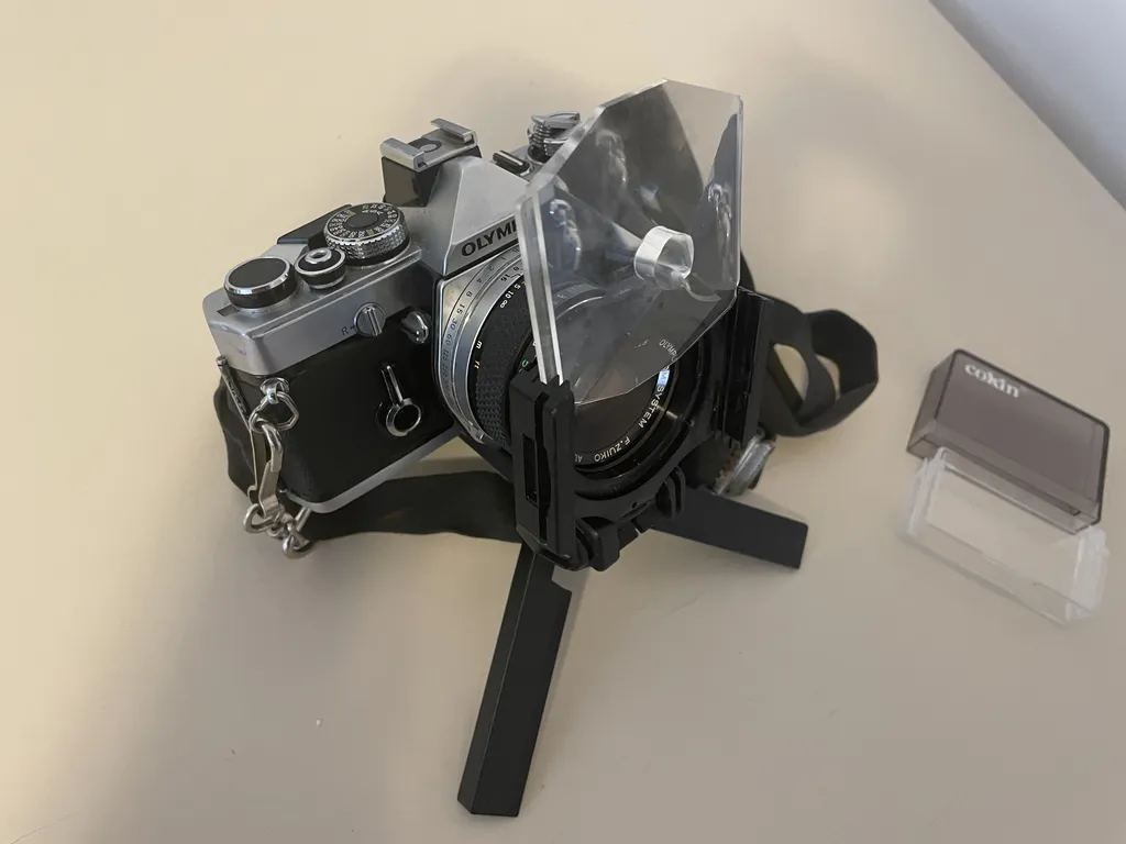 a black and silver metal camera on a tiny tripod. On the front of the lens is a black piece of plastic with ridged slots on either side. a thick, angular piece of plastic with a hole in the center is partially slotted in