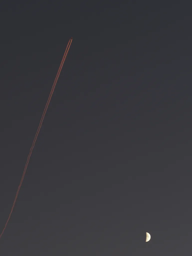 a contrail lit up by a setting sun by a quarter moon